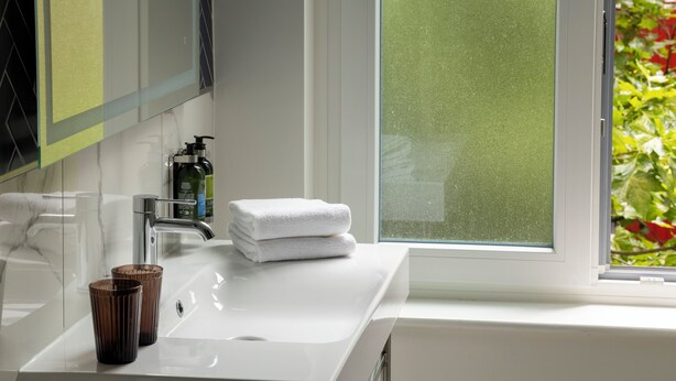 A bathroom with a white sink, two brown glasses, a stack of white towels, and a window with a view of greenery. - The Augustin (Brussels)