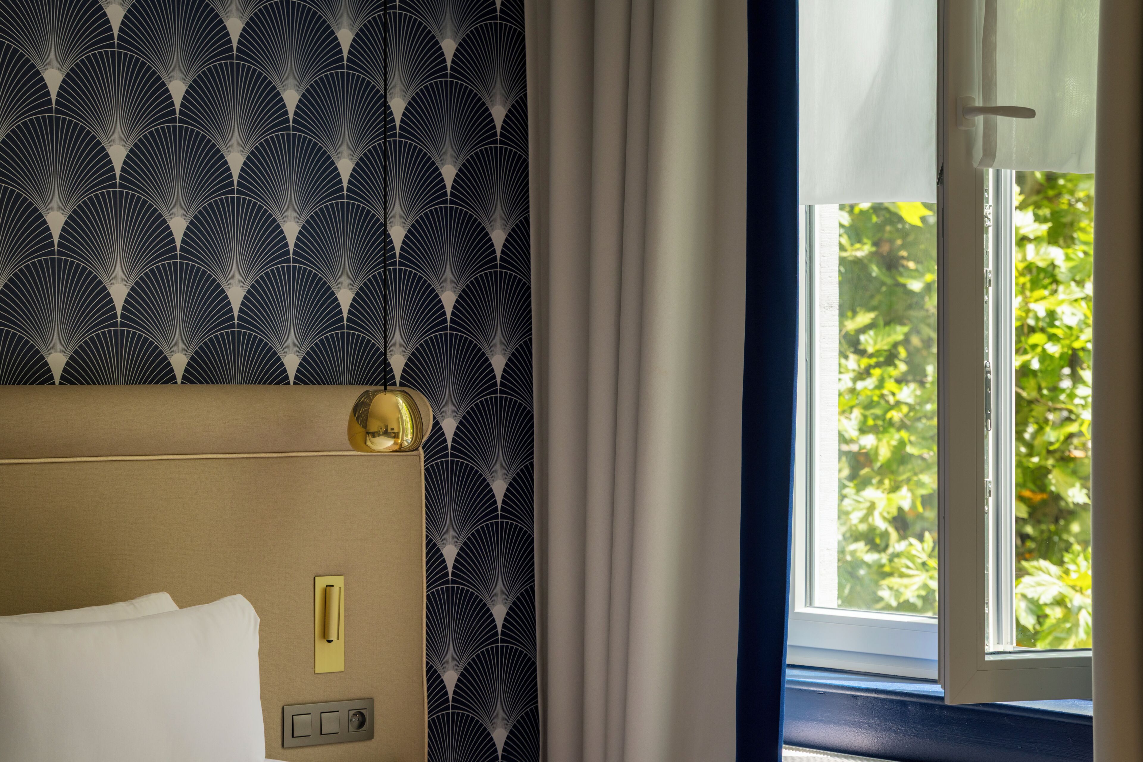 Habitación de hotel con papel tapiz estampado, una cama con ropa de cama blanca, una mesita de noche con lámpara y una ventana con cortinas.