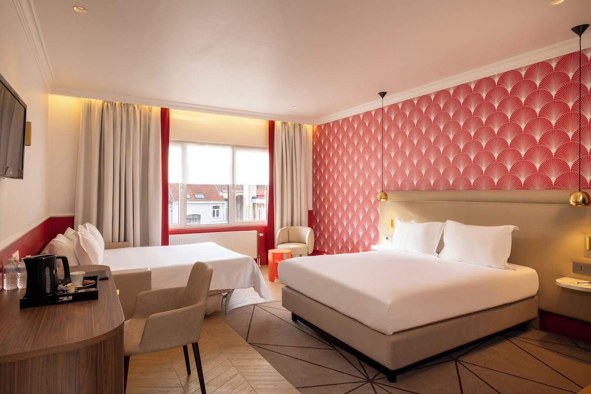 Une chambre d’hôtel avec deux lits, un bureau et une chaise. Il y a une fenêtre avec des rideaux, un mur à motifs rouges et une vue sur les bâtiments à l’extérieur.