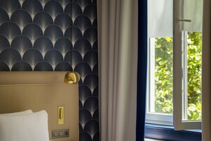Et hotelværelse med tapet med mønster, en seng med hvide sengetæpper, et natbord med lampe og et vindue med gardiner.
