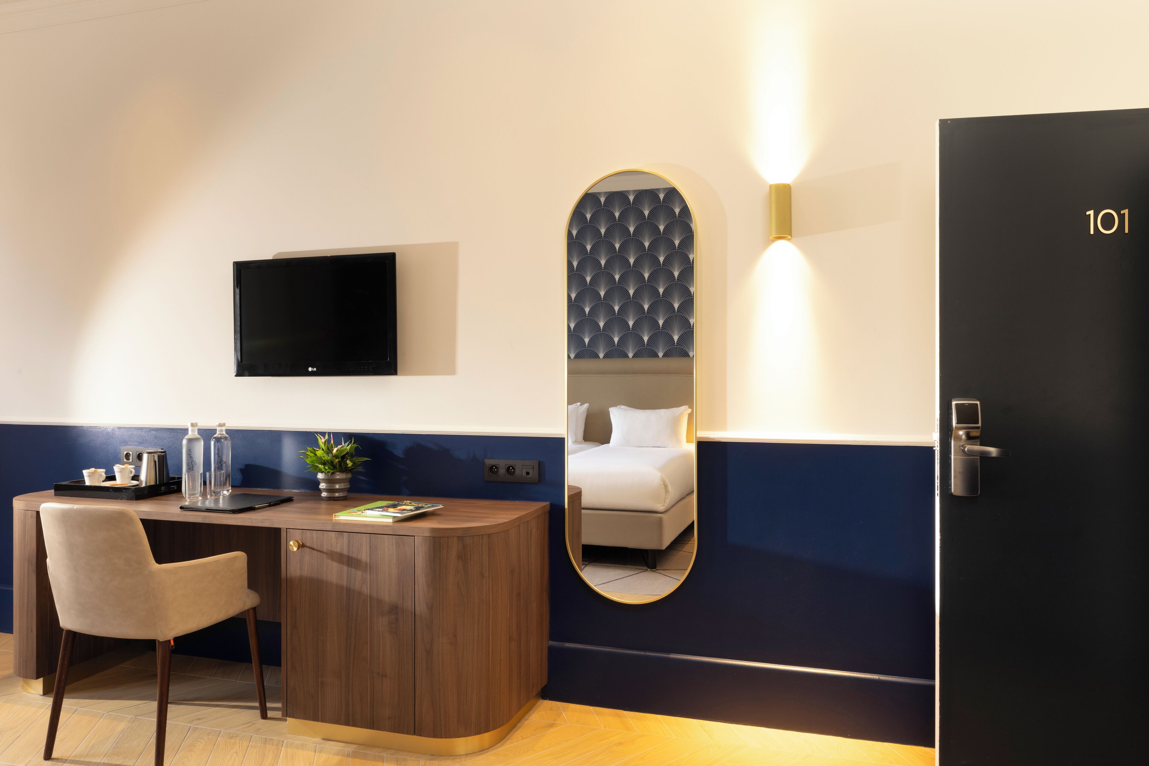 Une chambre d’hôtel équipée d’un bureau, d’une chaise, d’une télévision, d’un miroir et d’une porte portant le numéro 101.