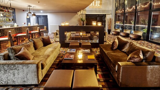 A modern lounge area with sofas, a coffee table, and a bar with bottles. - The Augustin (Brussels)