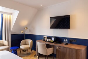 A hotel room with a desk, two chairs, a TV, a coffee maker, and a small table with a blue base.