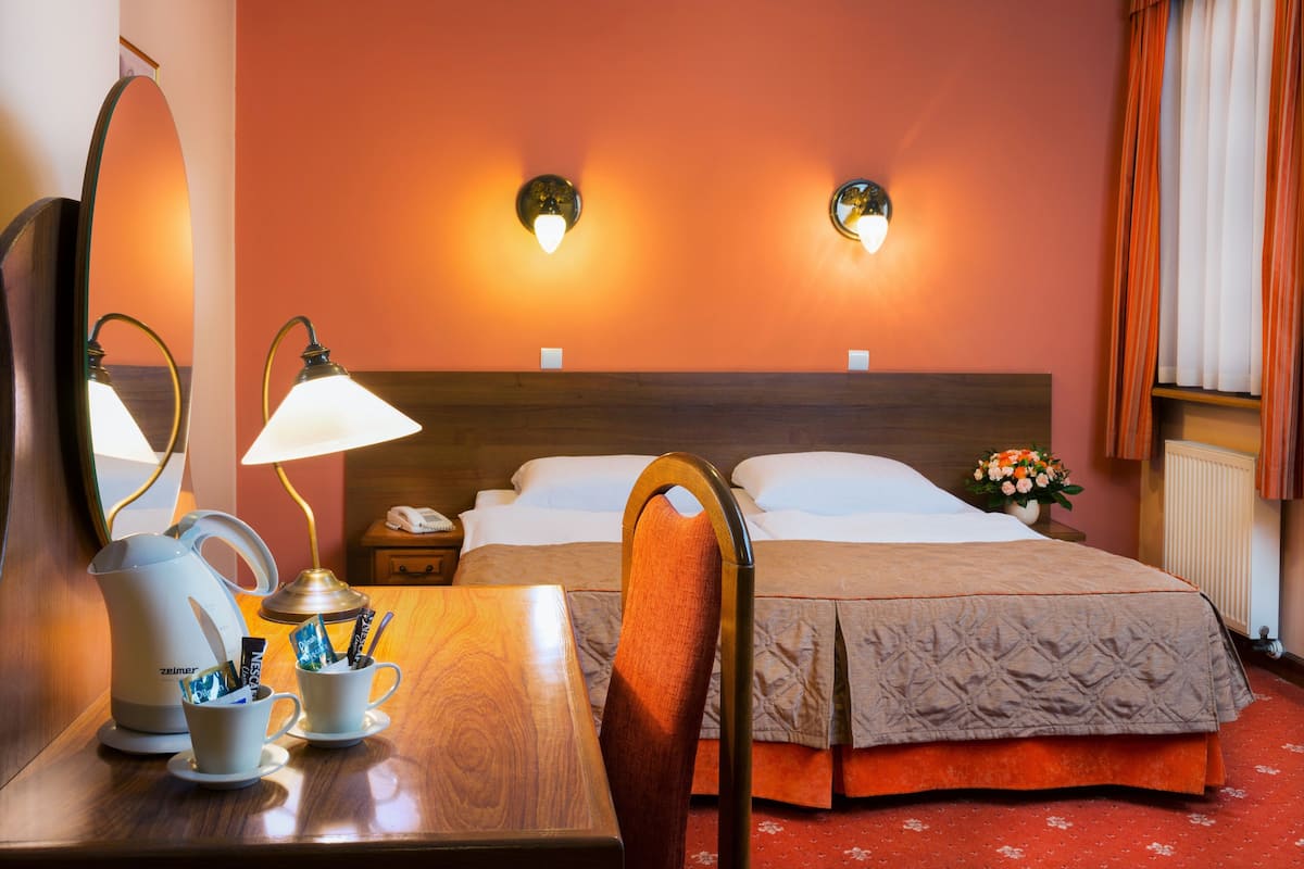 A hotel room with a wooden desk, a bed with a brown headboard, two wall-mounted lamps, a mirror, a small table with a telephone and a vase of flowers.