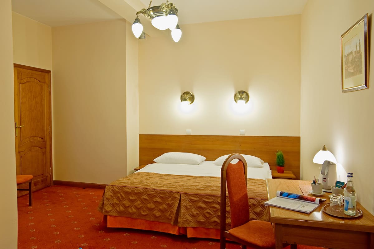 A hotel room with a bed, a desk with a lamp, a chair, a small table with a bottle, and a framed picture on the wall.