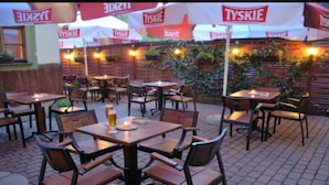 An outdoor dining area with wooden tables and chairs, covered by umbrellas with the Tyskie logo, and a brick wall with greenery in the background. - Hotel Regent (Kraków)