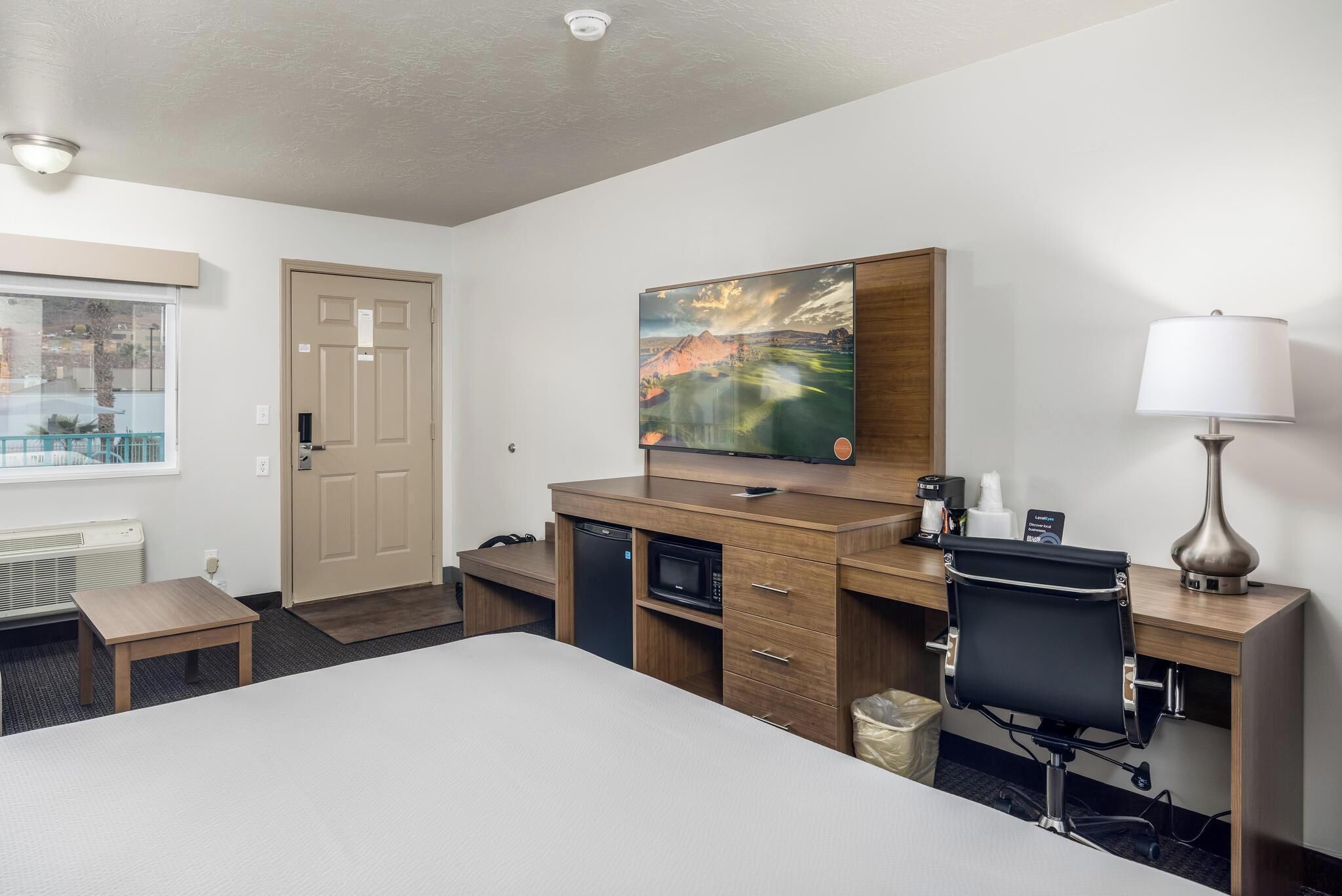 A hotel room with a bed, desk, chair, microwave, and a painting on the wall.