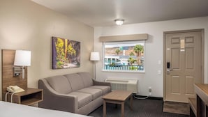 A hotel room with a bed, a sofa, a small table, a lamp, a painting, a window with a view of a building and palm trees, and a door. - St. George Inn & Suites (St. George)