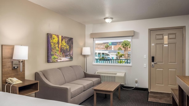 A hotel room with a bed, a sofa, a small table, a lamp, a painting, a window with a view of a building and palm trees, and a door. - St. George Inn & Suites (St. George)