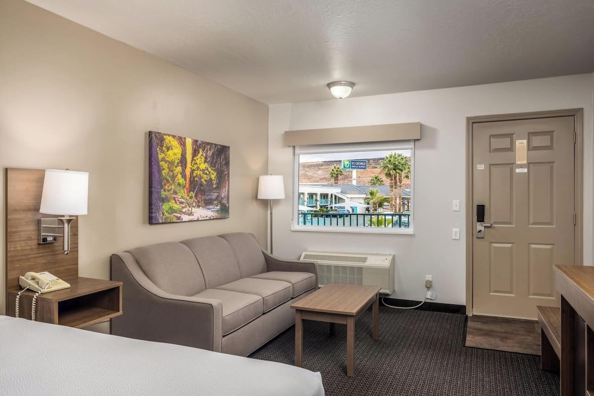 A hotel room with a bed, a sofa, a small table, a lamp, a painting, a window with a view of a building and palm trees, and a door.