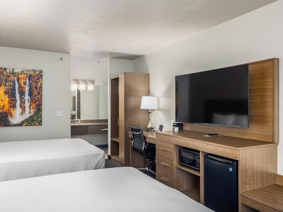 Habitación de hotel con dos camas, televisión, escritorio con lámpara, refrigerador pequeño y un baño visible al fondo.