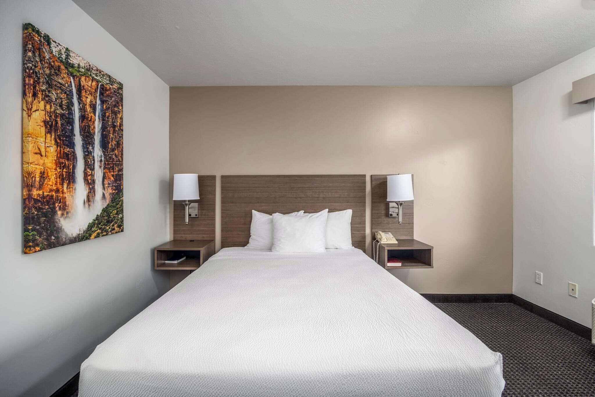 A hotel room with a large bed, two bedside tables with lamps, a painting on the wall, and a plain carpeted floor.
