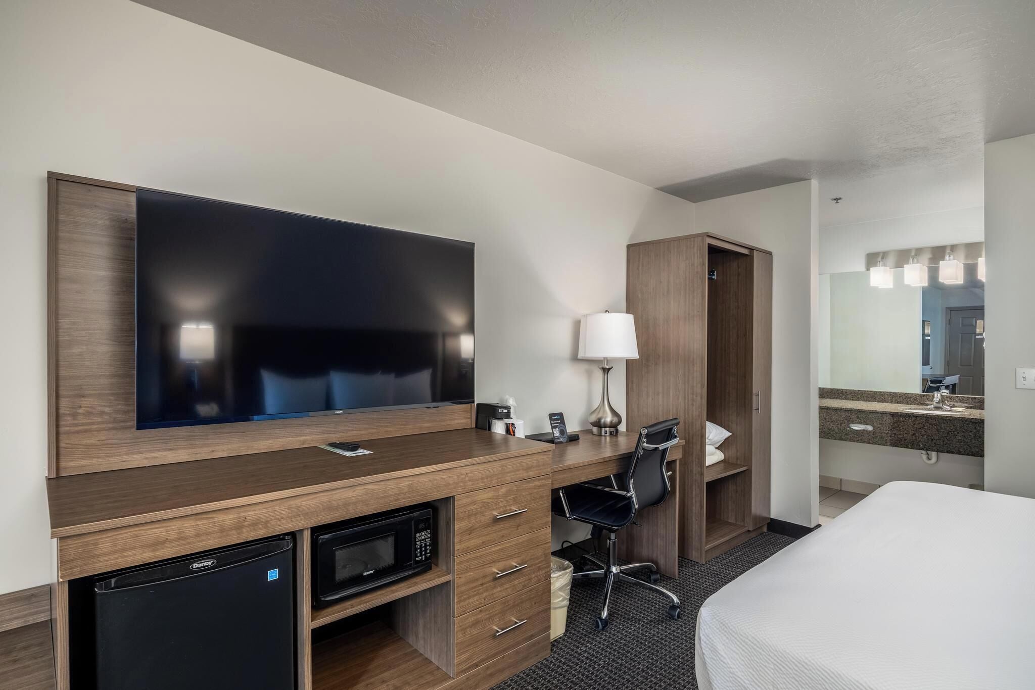 A hotel room with a flat-screen TV, a desk with a lamp, a microwave, a mini-fridge, a black office chair, a bed, a wooden wardrobe, and a bathroom with a sink and mirror.