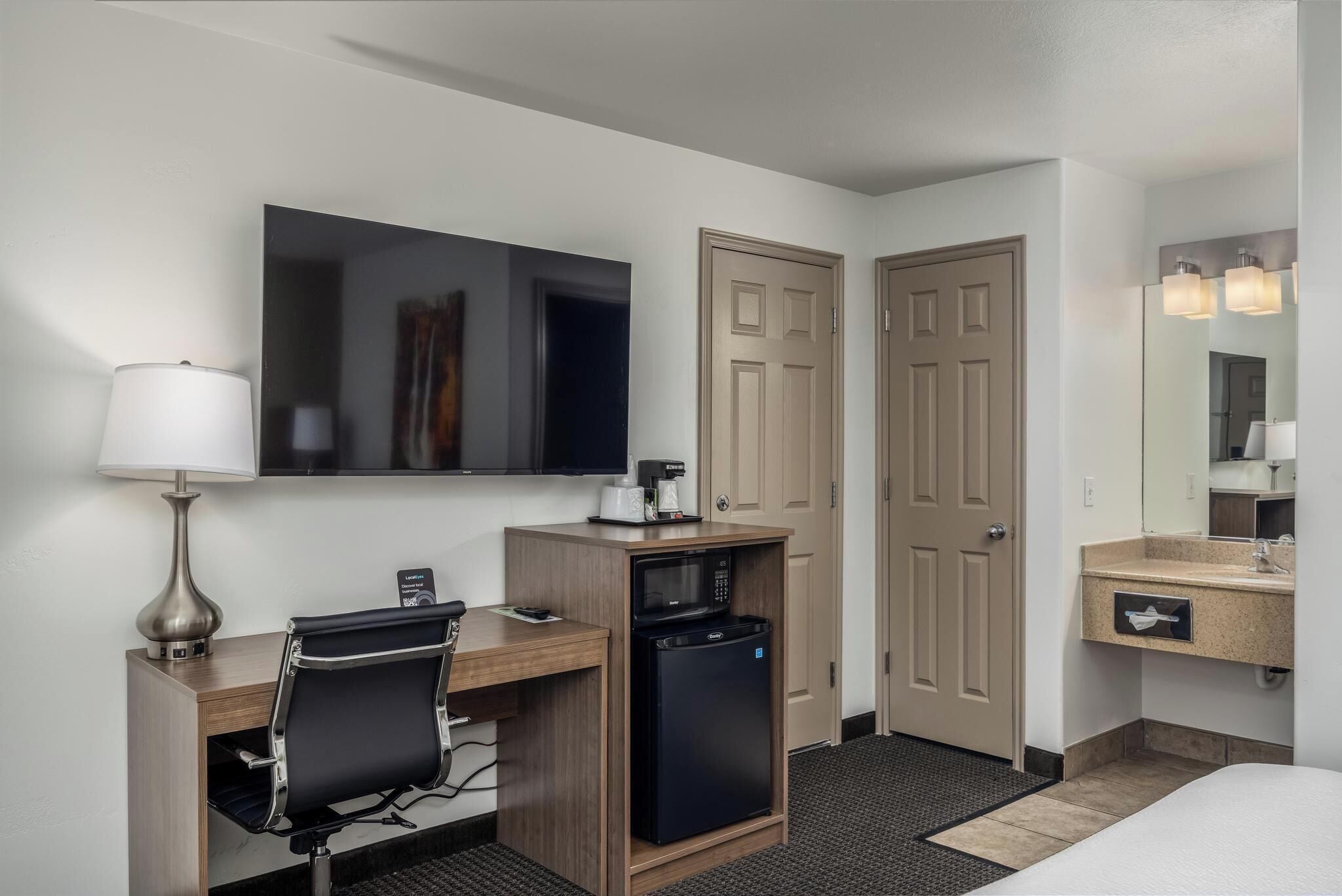 A hotel room with a desk, a black office chair, a microwave, a coffee maker, a TV, a mirror, and a bathroom with a sink and mirror.