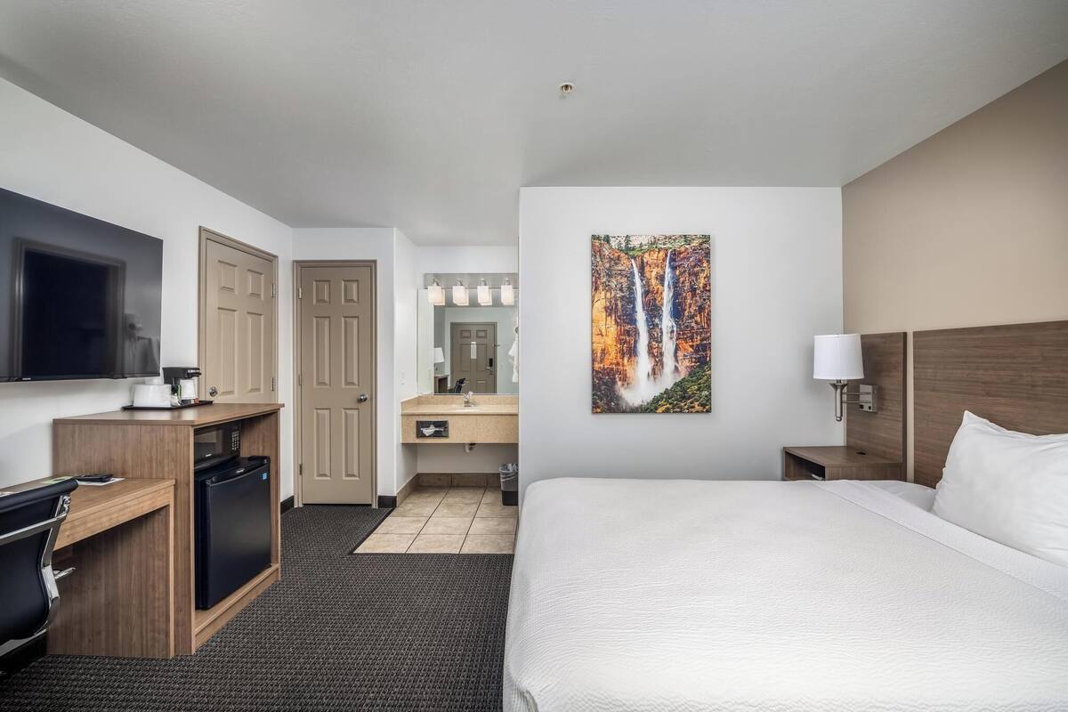 A hotel room with a bed, a desk with a coffee maker, a TV, a bathroom with a sink and mirror, and a painting of a waterfall.