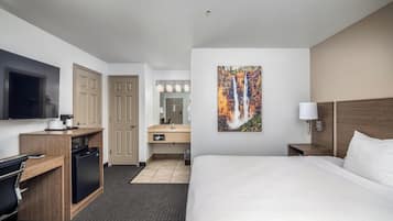 A hotel room with a bed, a desk with a coffee maker, a TV, a bathroom with a sink and mirror, and a painting of a waterfall.