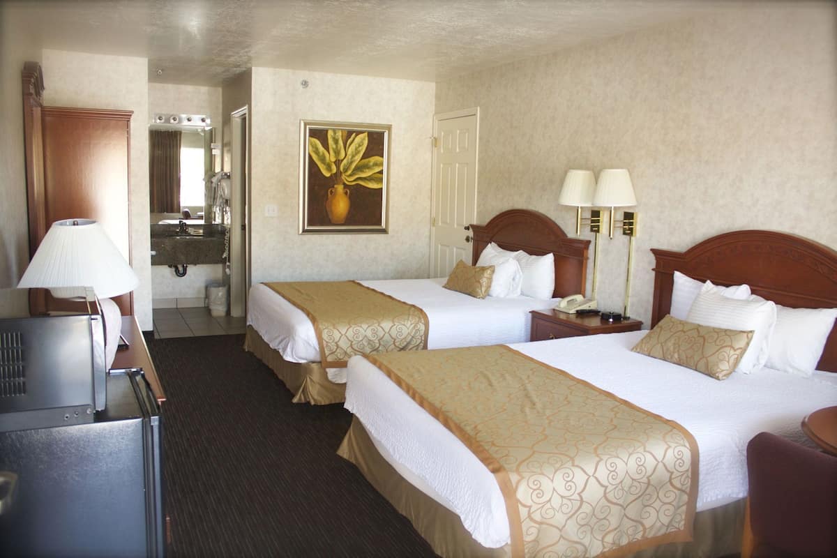 A hotel room with two beds, a desk, a lamp, a mirror, a painting, and a bathroom.