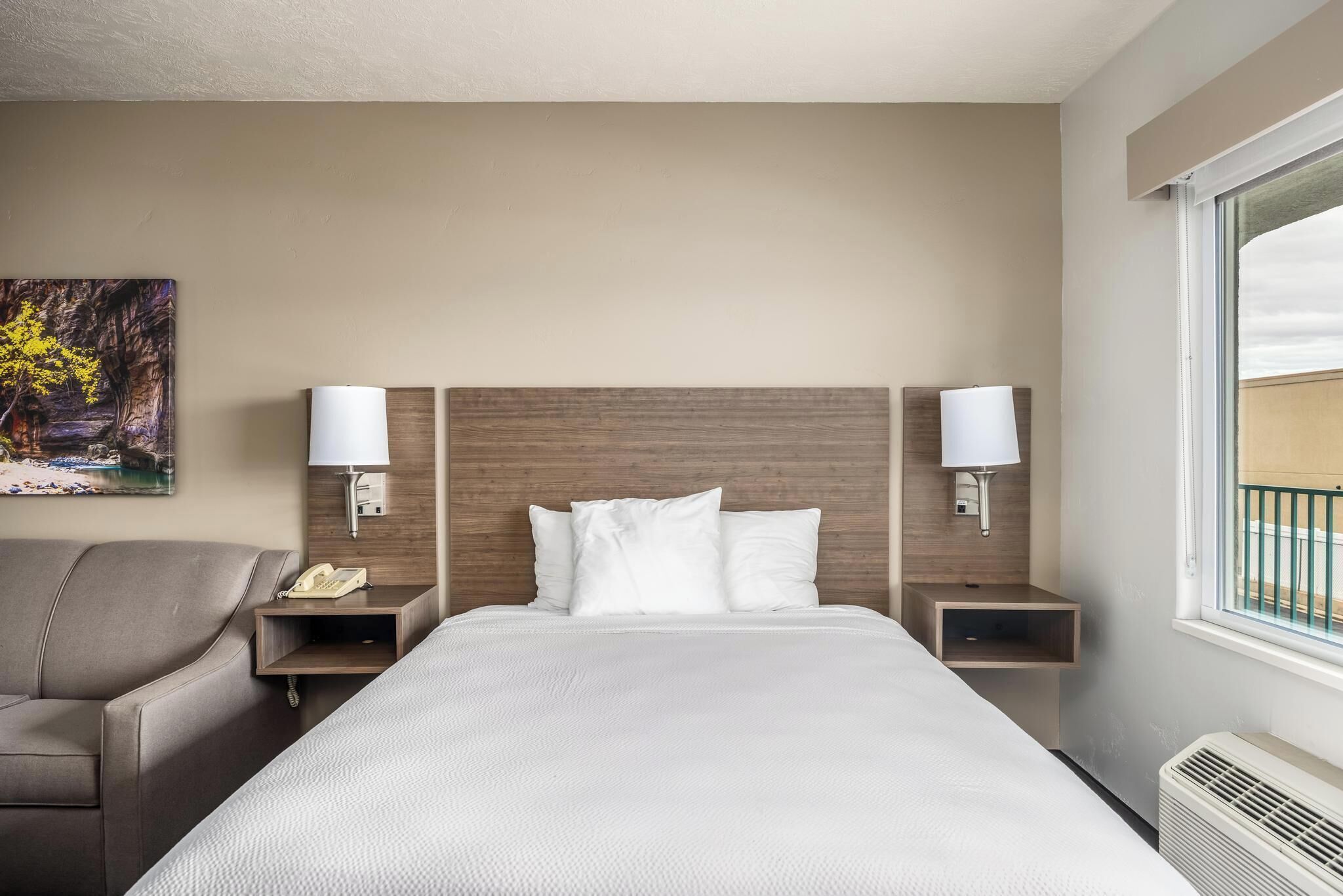 Habitación de hotel con una cama grande, dos mesitas de noche, una lámpara, un teléfono, un cuadro y una ventana con vistas.