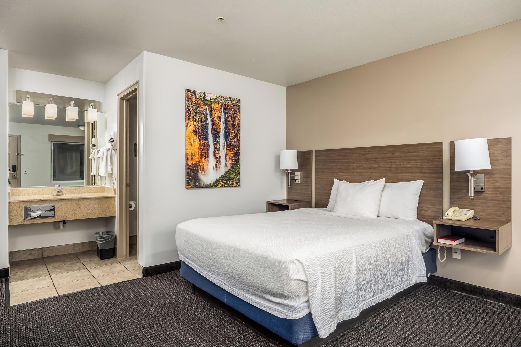 A hotel room with a large bed, a bathroom with a mirror and lights, and a painting on the wall.