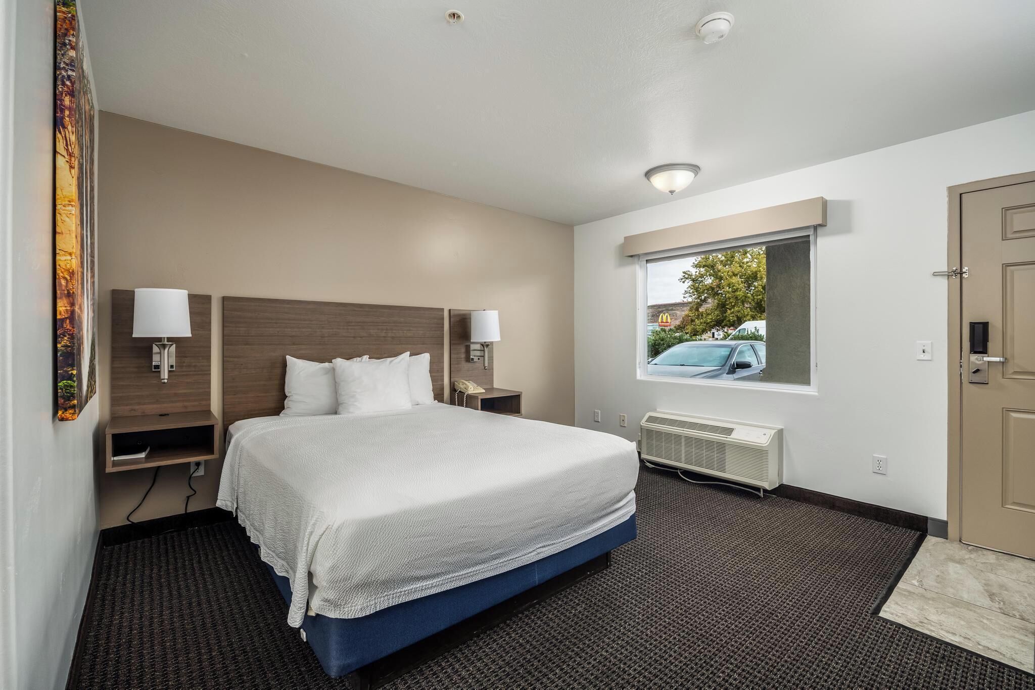 A hotel room with a large bed, bedside lamps, a window with a view of a car, and a door.