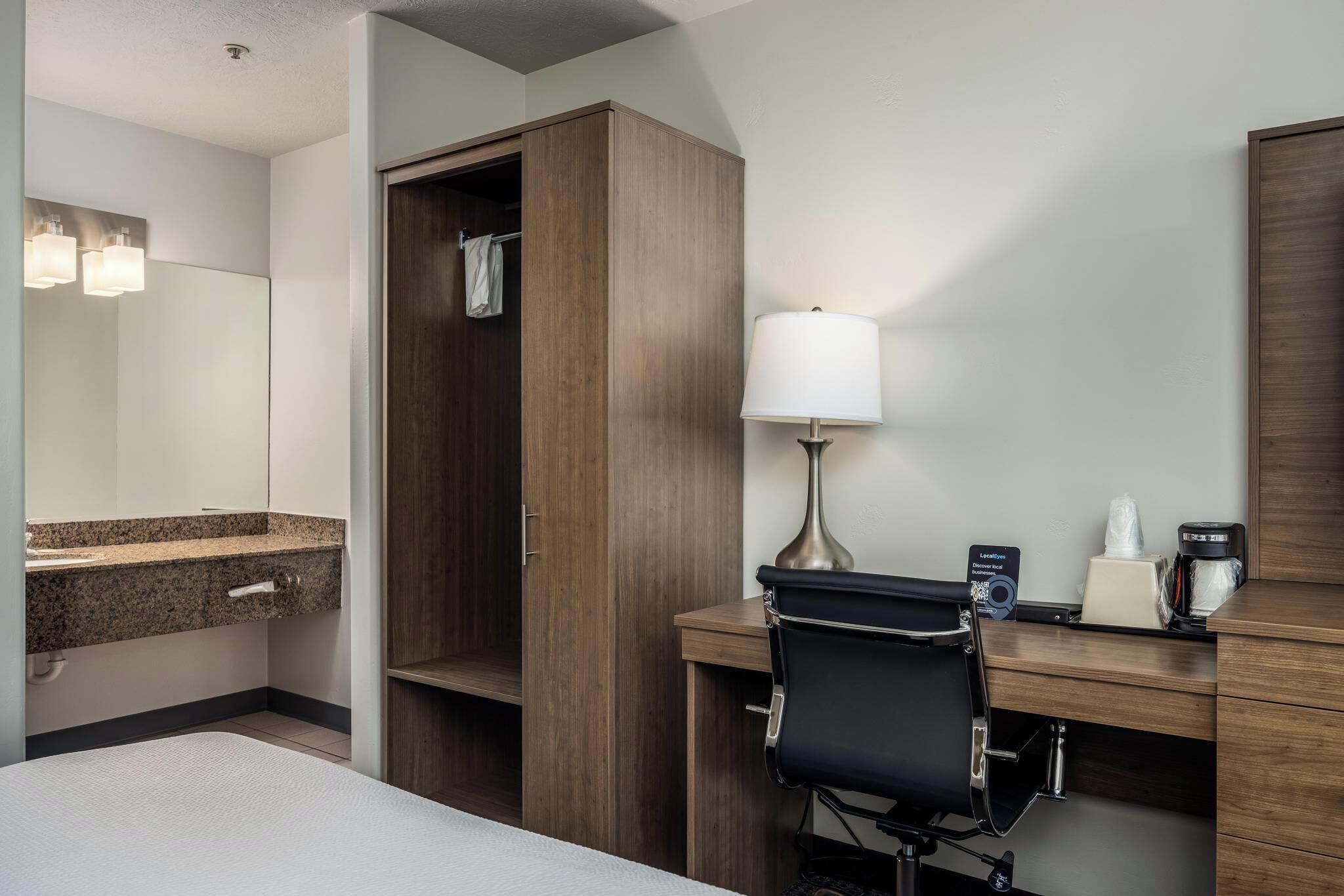 Habitación de hotel con una cama, un escritorio con silla, armario de madera, espejo, lámpara y cafetera.