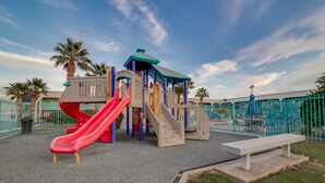 A playground with slides and climbing structures, surrounded by a fence and palm trees. - St. George Inn & Suites (St. George)