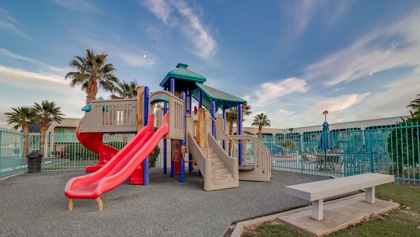 A playground with slides and climbing structures, surrounded by a fence and palm trees. - St. George Inn & Suites (St. George)