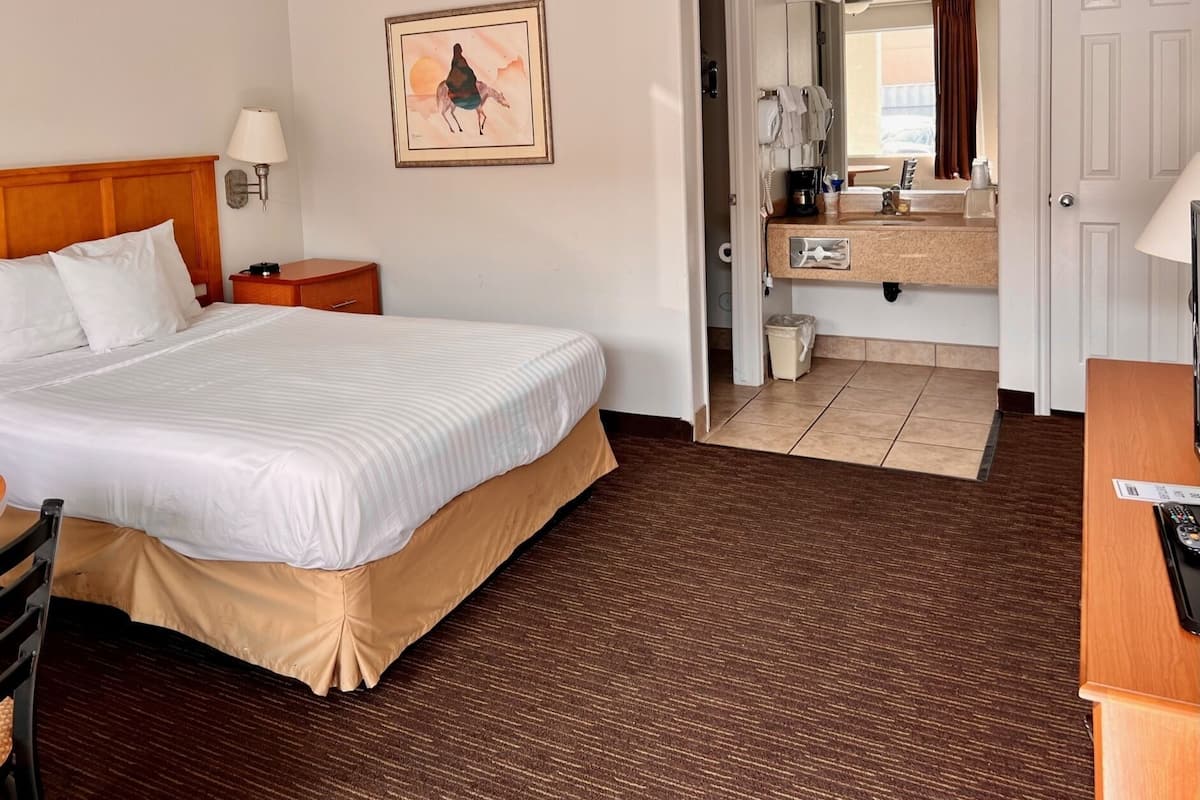 A hotel room with a bed, a nightstand, a painting, a bathroom with a sink, mirror, and towel rack.
