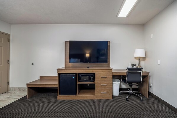 A hotel room with a wooden desk, a television, a microwave, a refrigerator, a chair, and a lamp.
