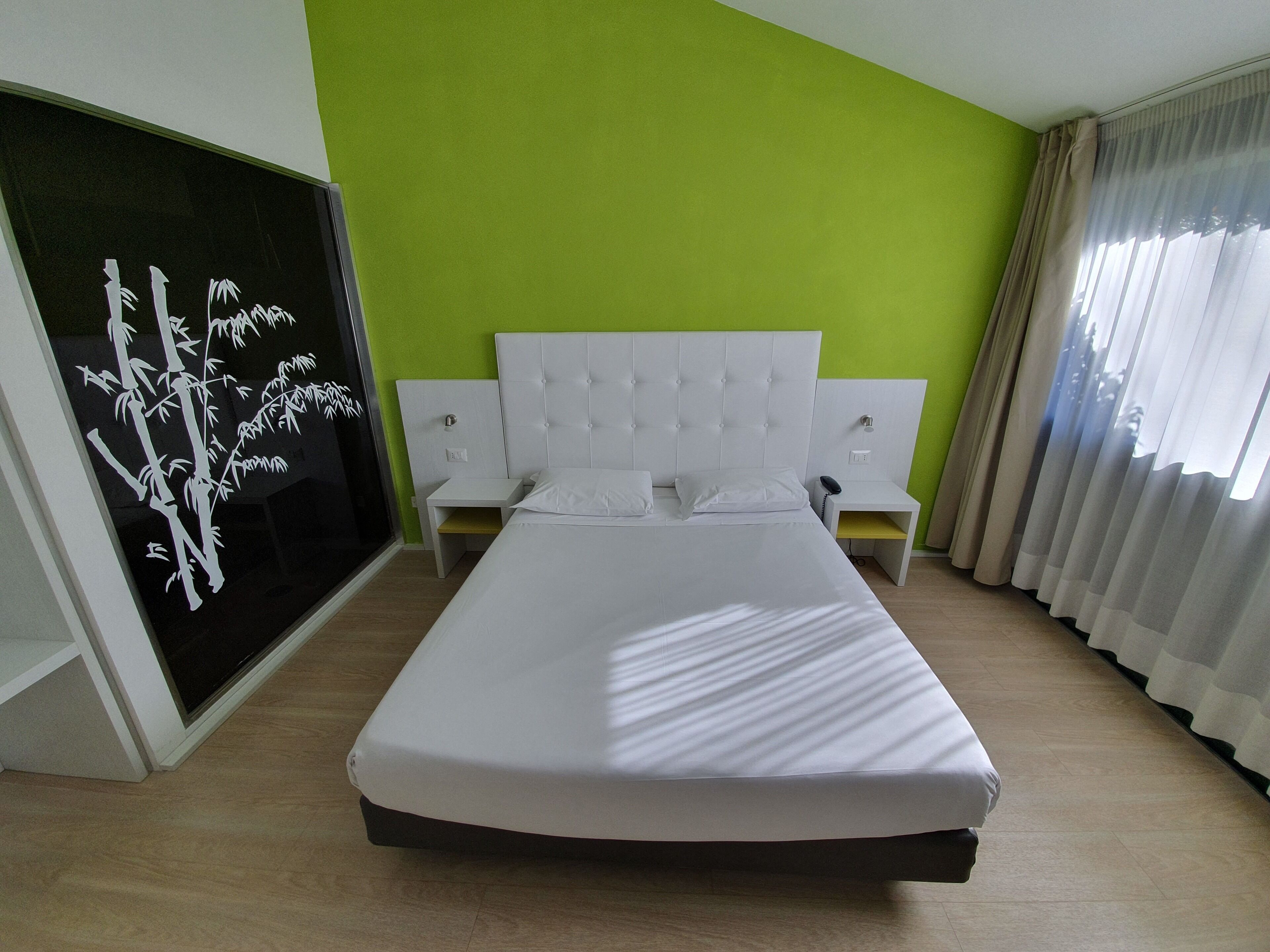 Una habitación de hotel ordenada con cama, mesitas de noche y un amplio ventanal con cortinas.
