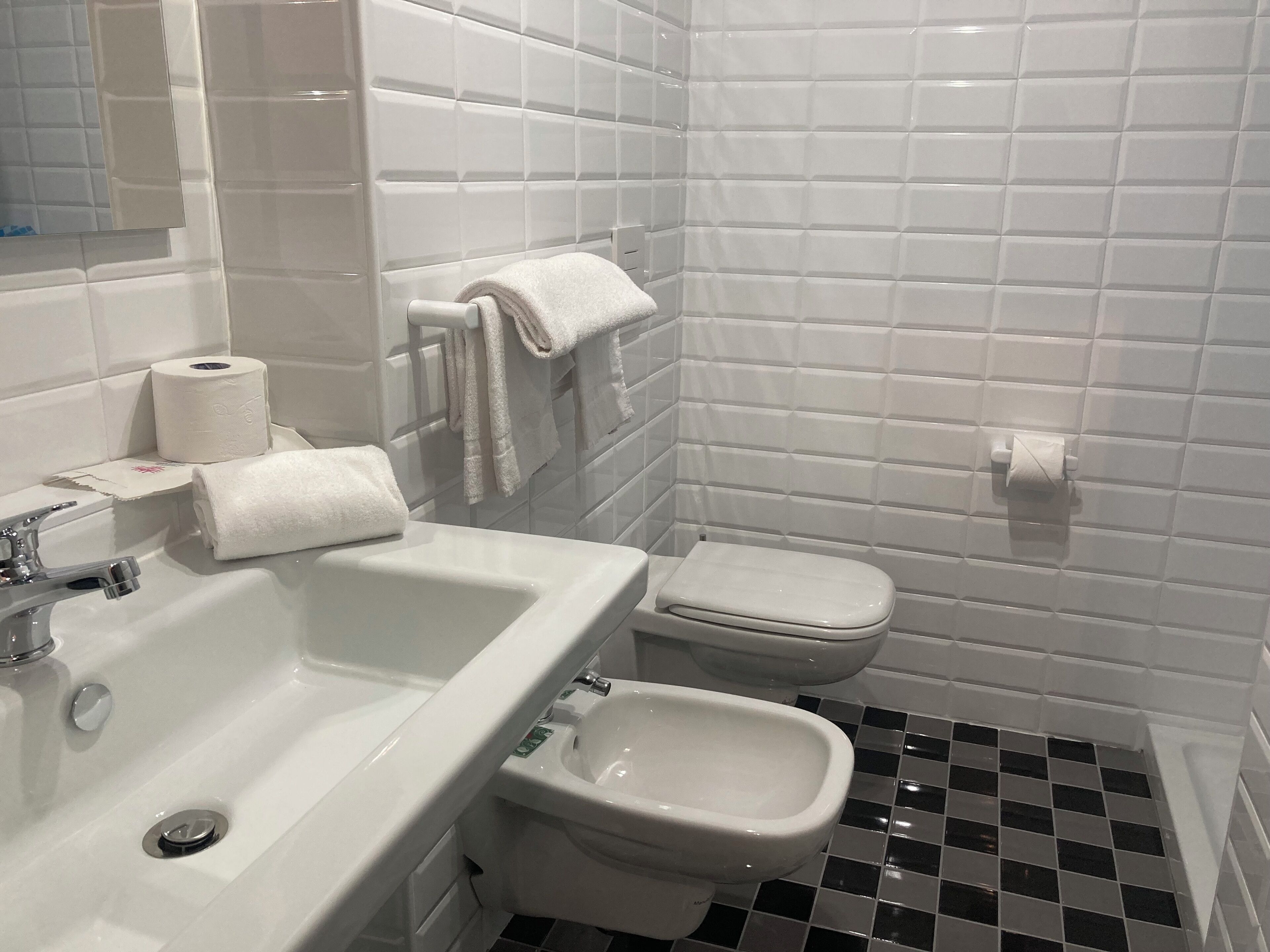 Un baño con lavamanos, inodoro y bidé blancos, con azulejos en blanco y negro.
