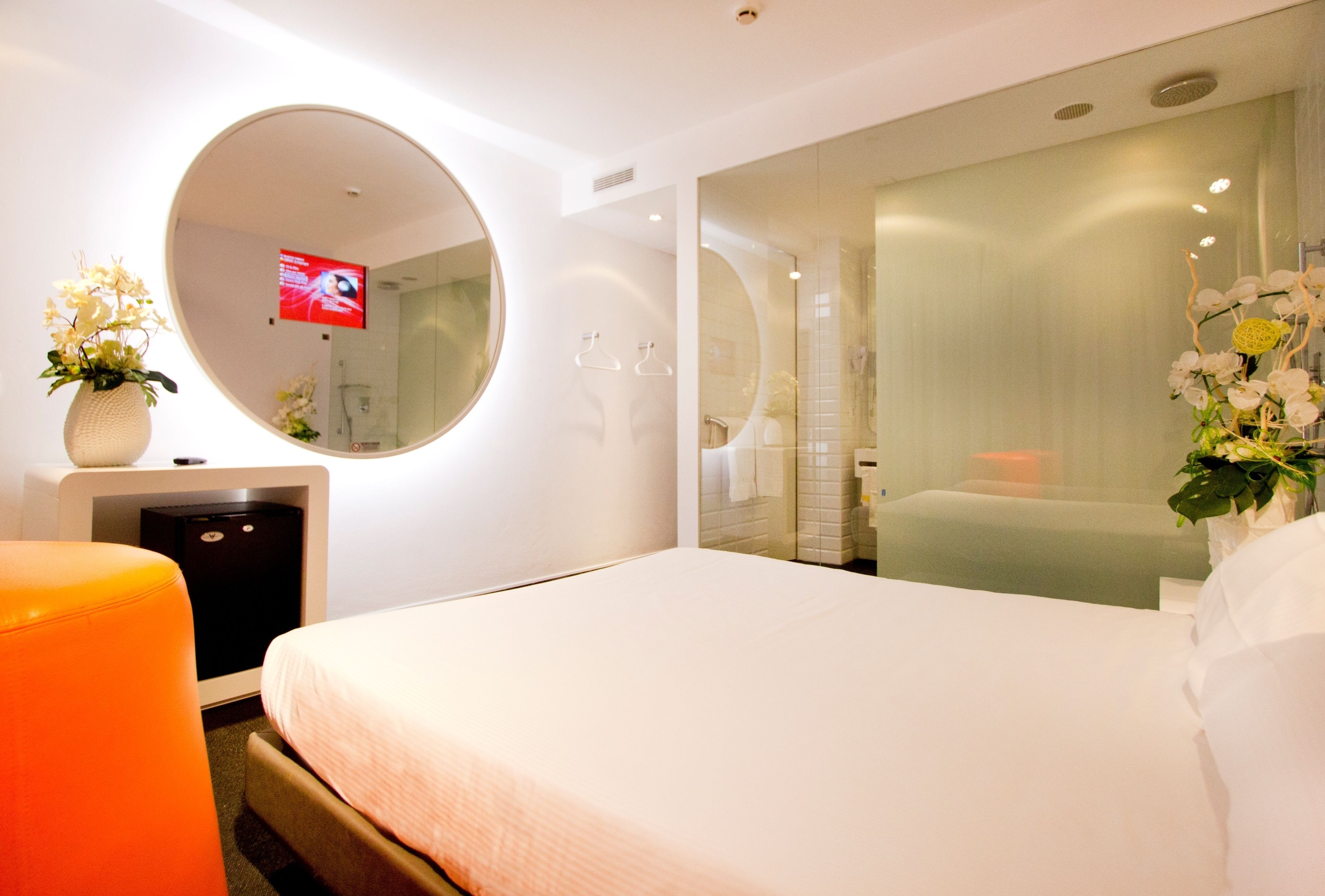 A modern hotel room with a large bed, a round mirror, a small desk with a lamp, and a TV.