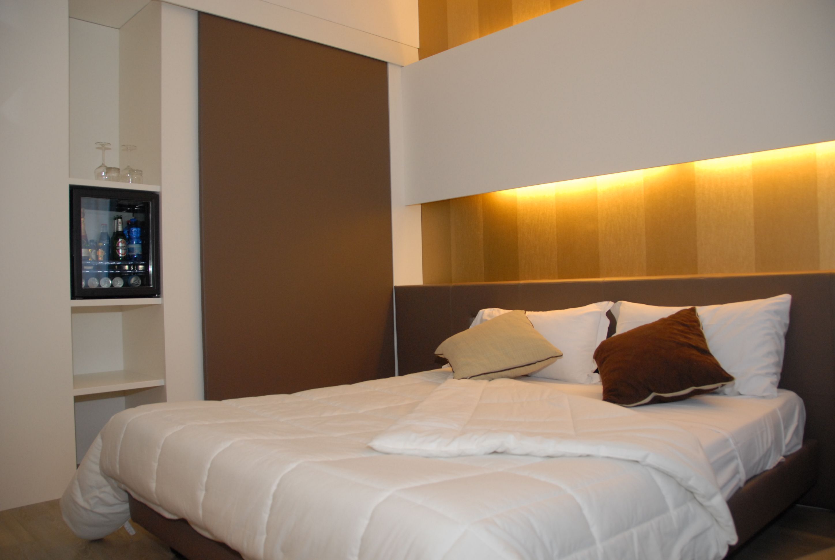 A neatly made bed with white bedding and two pillows, a built-in shelf with bottles and glasses, and a wall-mounted light fixture above the bed.