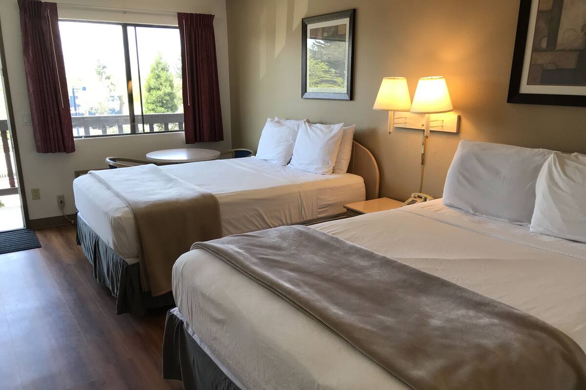 A hotel room with two beds, a small table, a chair, a lamp, and a window with curtains.