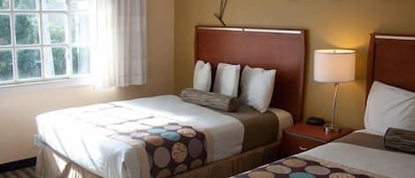 A hotel room with two beds, a window with curtains, a wall-mounted lamp, and a decorative plant.