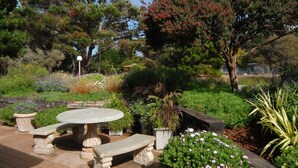 A garden patio with a round table and two benches, surrounded by various plants and trees. - Deer Haven Inn (Pacific Grove)