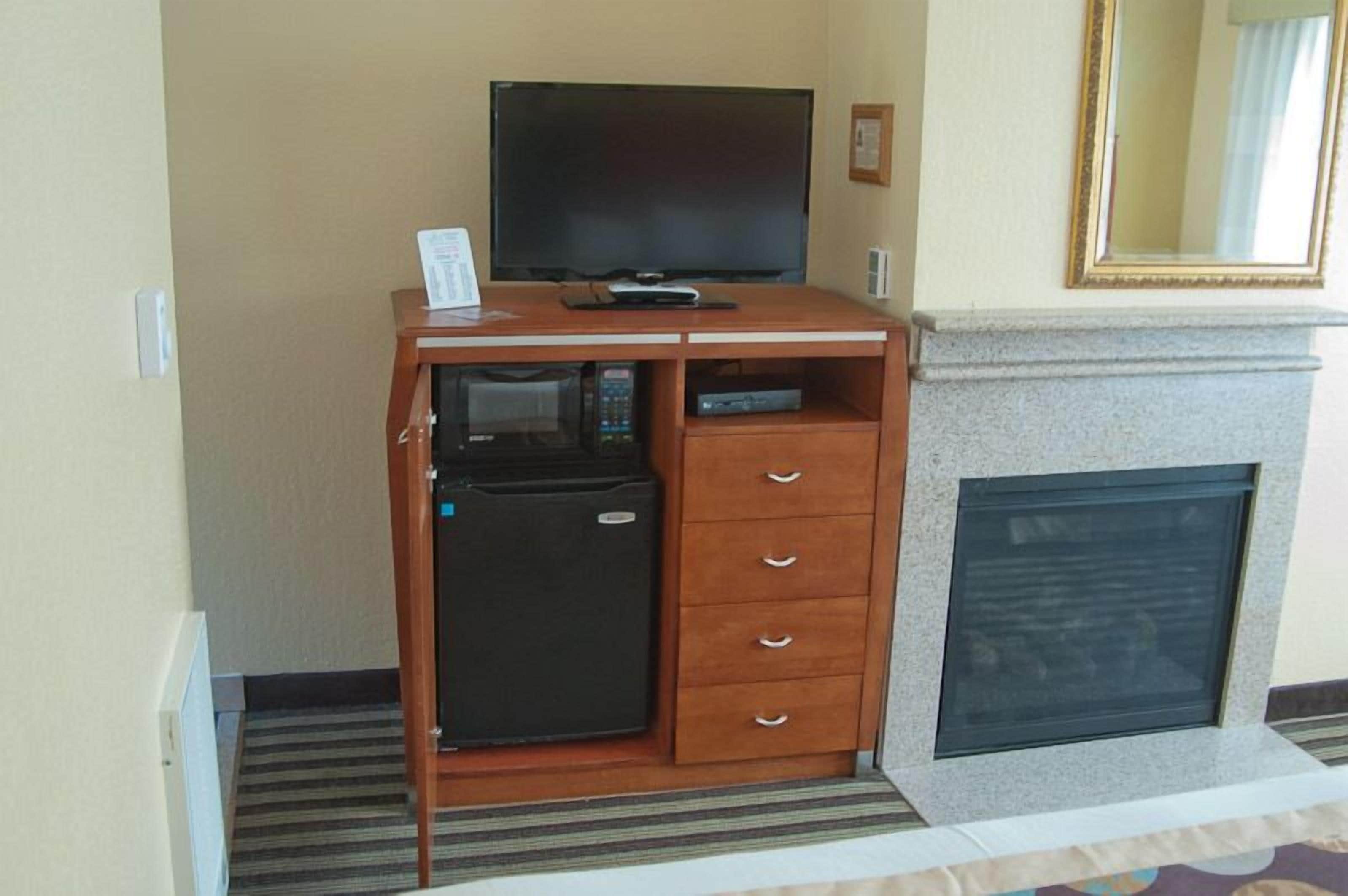 A compact hotel room with a flat-screen TV on a wooden cabinet, a small refrigerator, a fireplace, and a mirror above it.