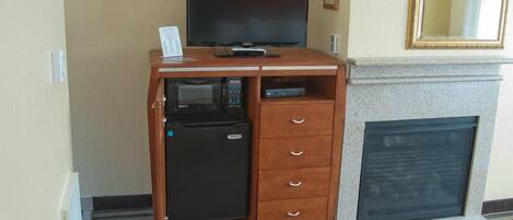 A compact hotel room with a flat-screen TV on a wooden cabinet, a small refrigerator, a fireplace, and a mirror above it.