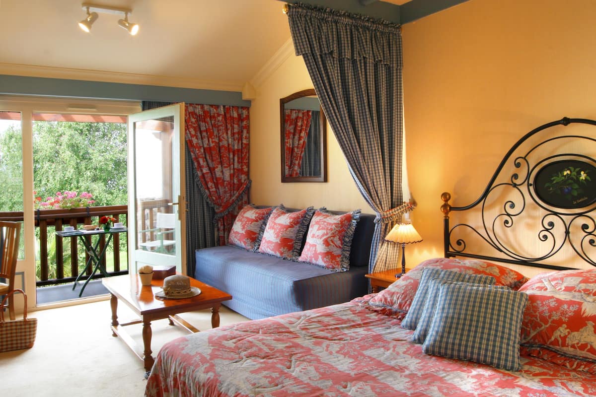 A bedroom with a bed, a sofa, a table, and a balcony with a view of greenery.