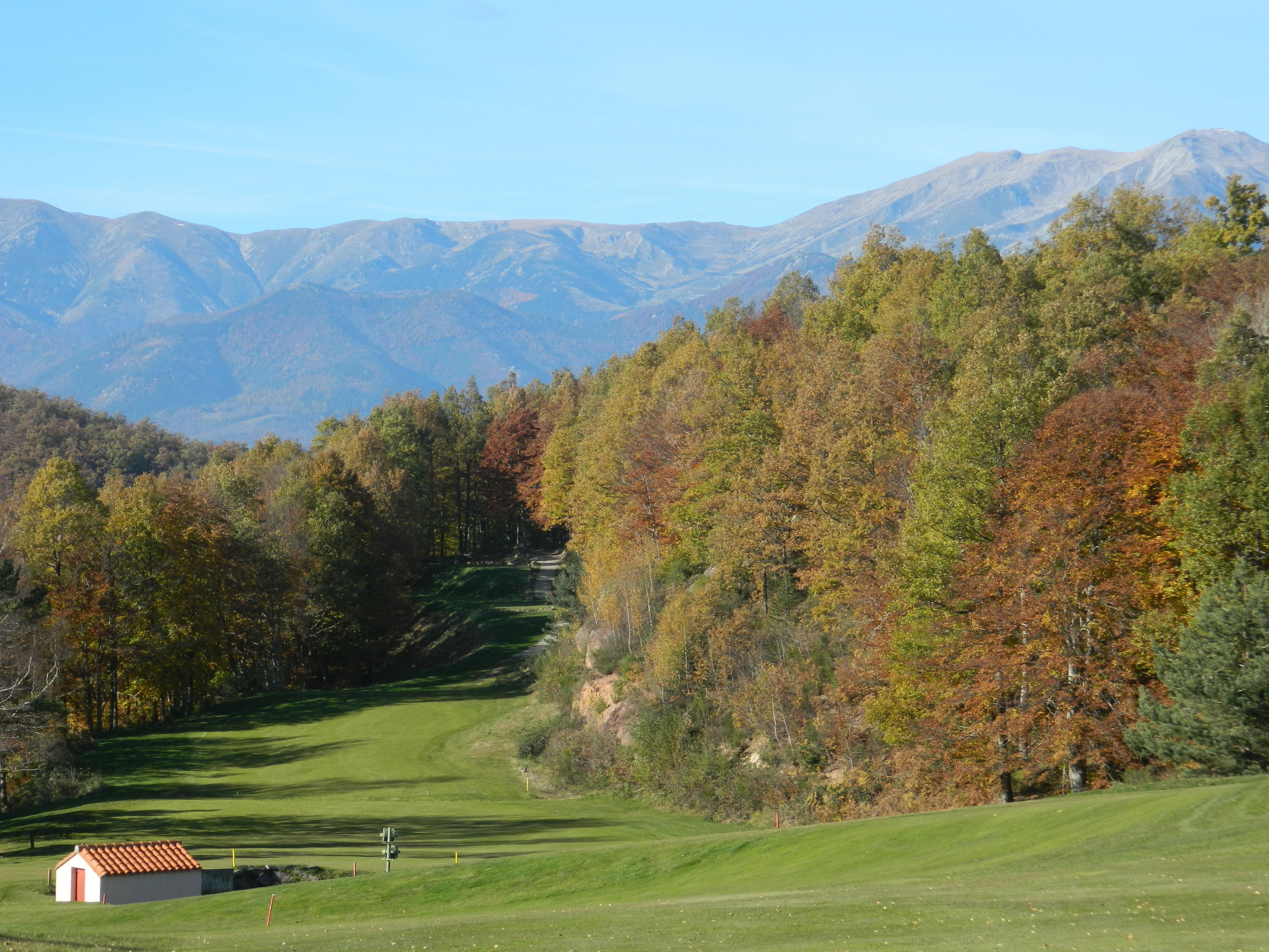 Un parcours de golf avec un petit bâtiment, entouré d’arbres aux couleurs automnales et avec des montagnes en arrière-plan.