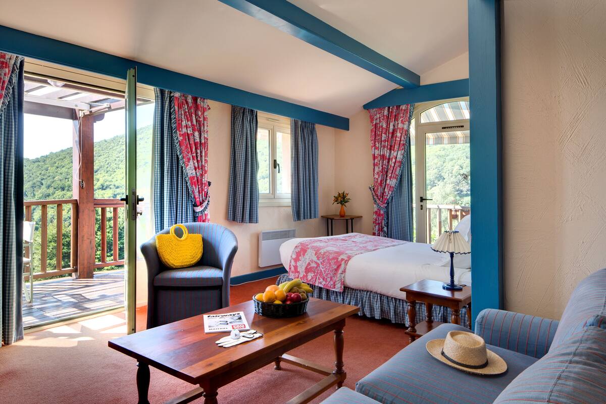 A bedroom with a bed, a chair, a table with a fruit basket, a lamp, and a view of a balcony with a wooden deck.