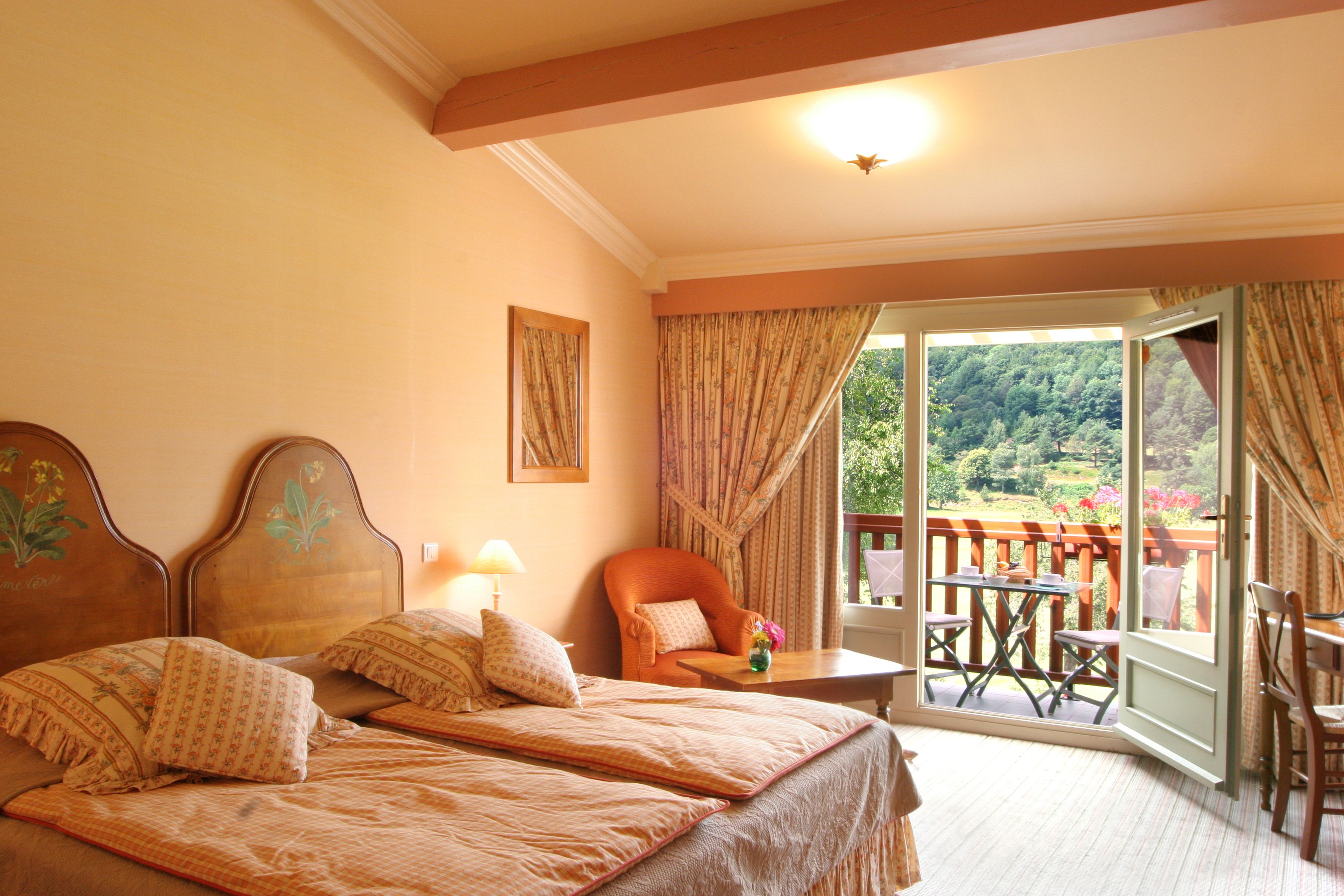 A bedroom with two beds, a chair, a table, and a balcony with a view of greenery.