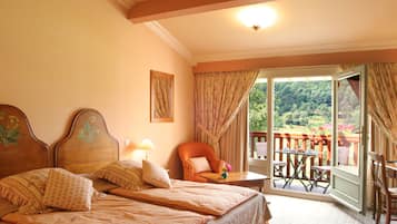 A bedroom with two beds, a chair, a table, and a balcony with a view of greenery.