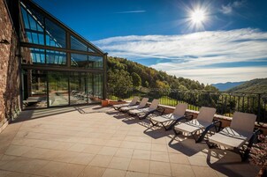 Terraço com espreguiçadeiras, um edifício de vidro e vista para um morro arborizado.