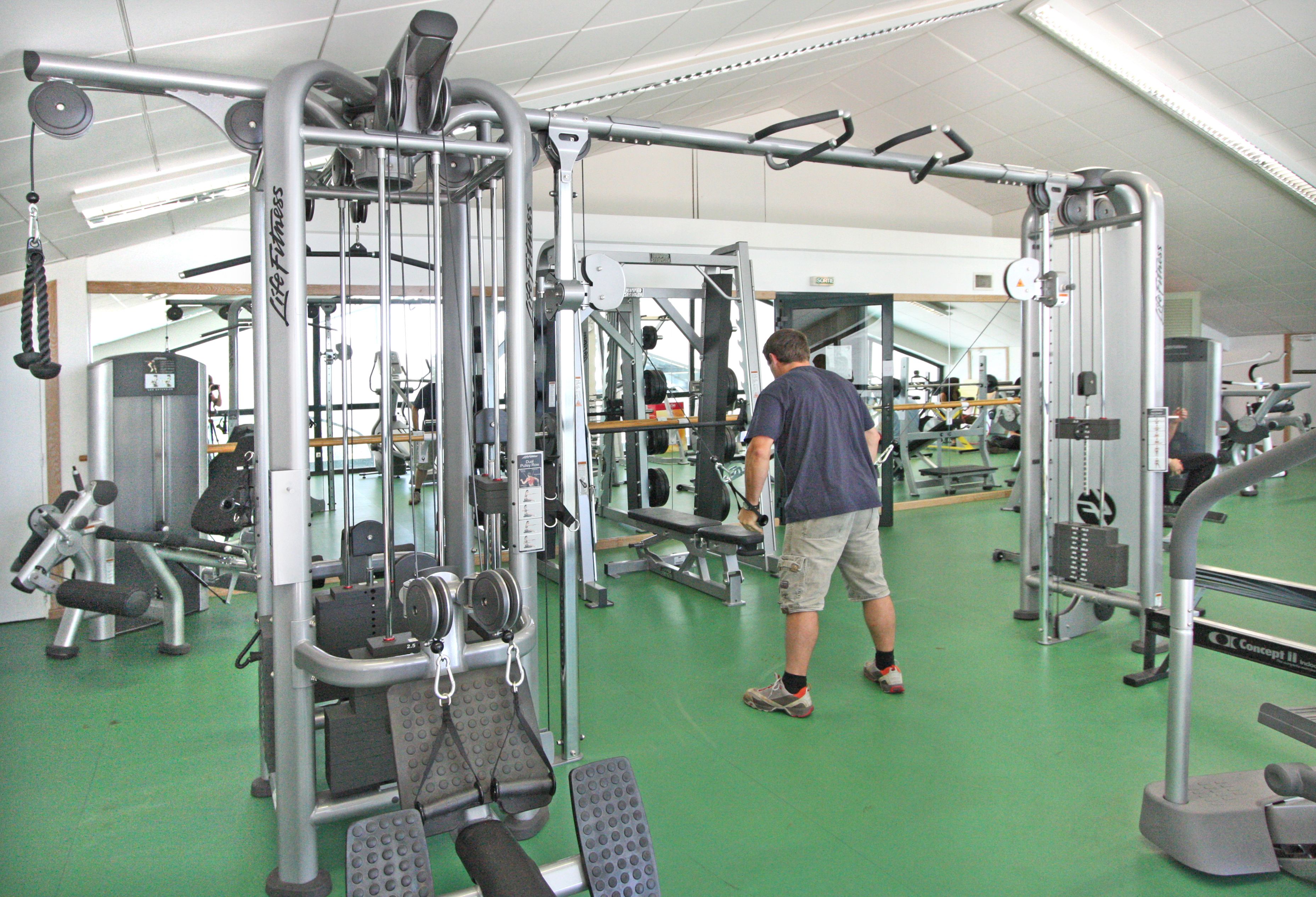 Une salle de sport équipée de divers appareils d’exercice, un homme utilisant un appareil à câbles et un sol vert.