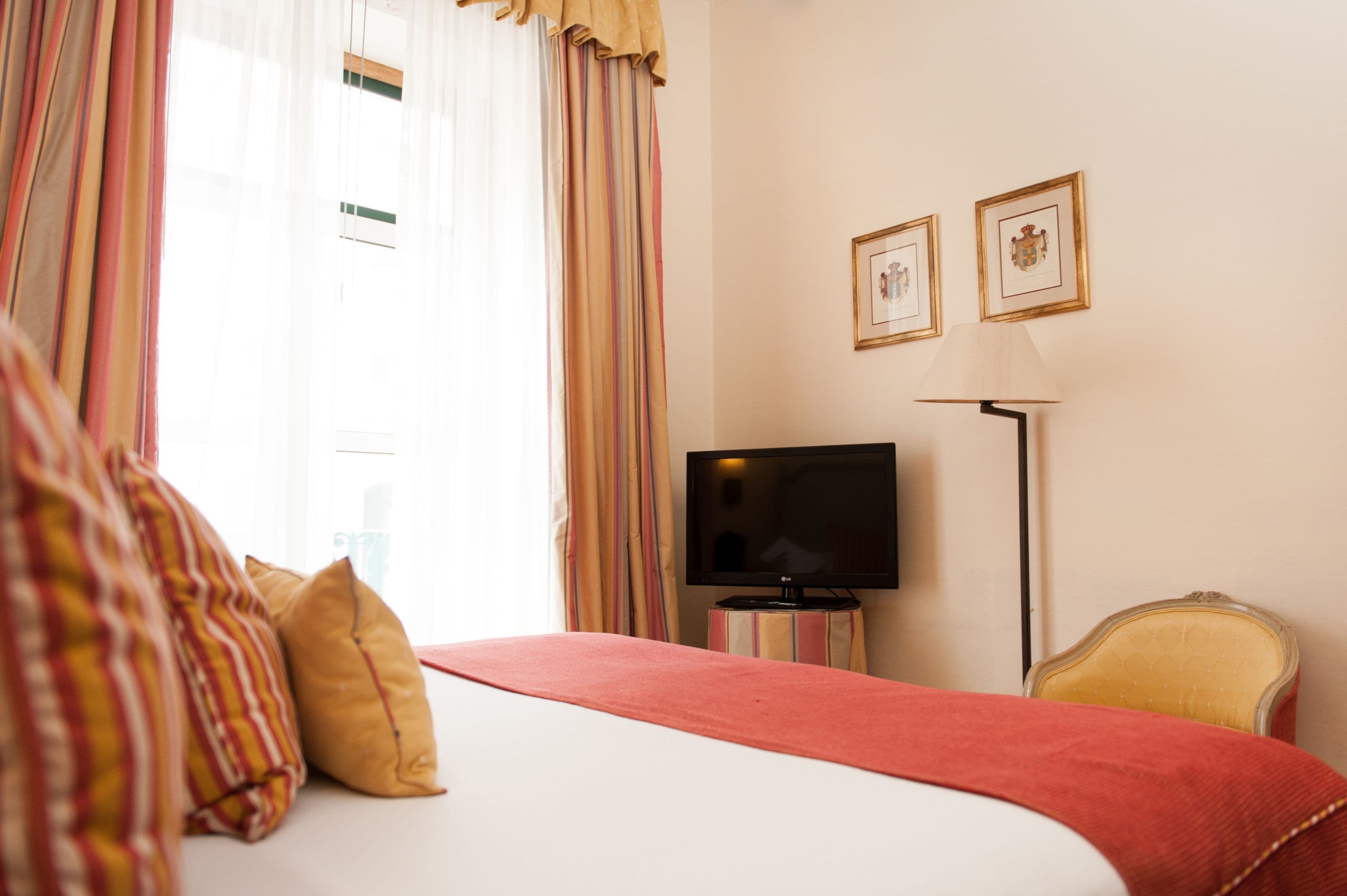 Habitación de hotel con cama, televisor sobre un soporte, una silla y dos cuadros en la pared.