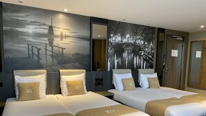 A hotel room with two beds, each with white linens and beige pillows, a large wall mural depicting a serene landscape, and a dark-colored carpet. - XO Hotels Infinity (Amsterdam)