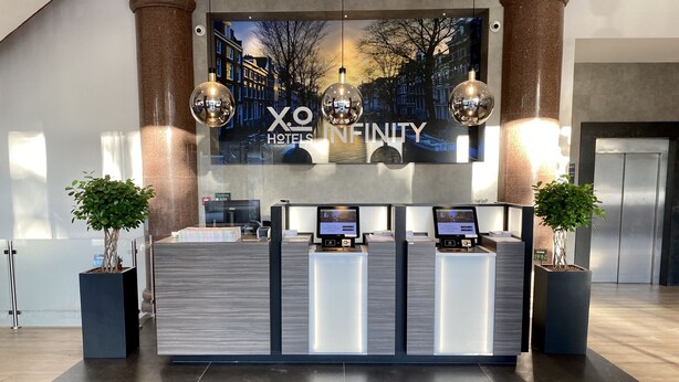 A hotel reception desk with a large sign reading "X.O Hotels Infinity" and two potted plants on either side. - XO Hotels Infinity (Amsterdam)