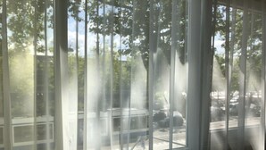 A view through a window with sheer curtains, revealing a parking lot and trees outside. - XO Hotels Infinity (Amsterdam)