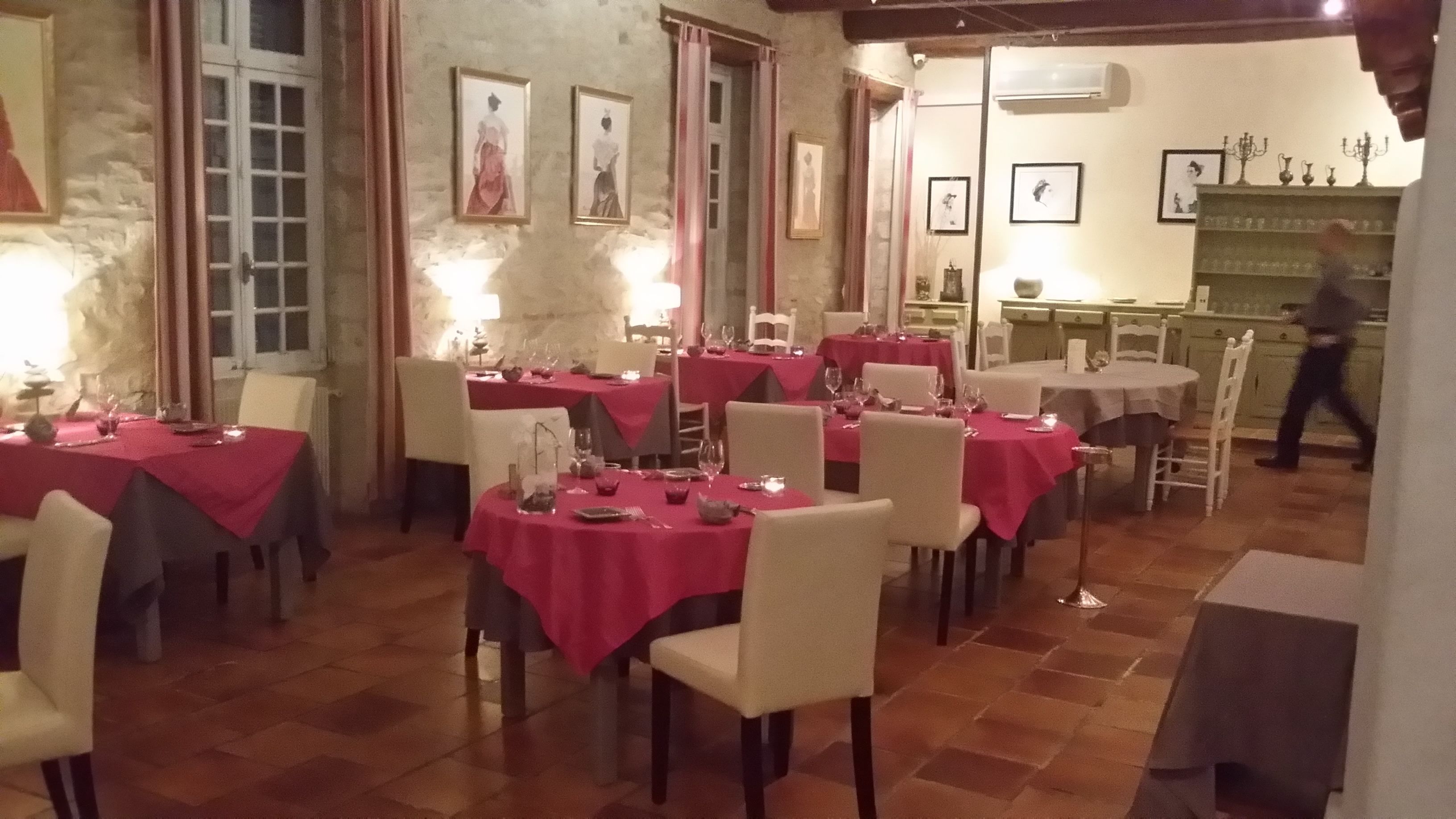 Un comedor con varias mesas redondas cubiertas con manteles rojos, sillas blancas y velas. Se observan cuadros en las paredes y una persona caminando al fondo.